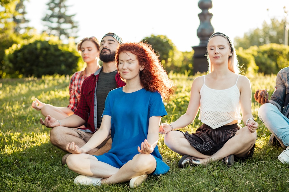 Group of adult friends meditating while practicing yoga outside in park.