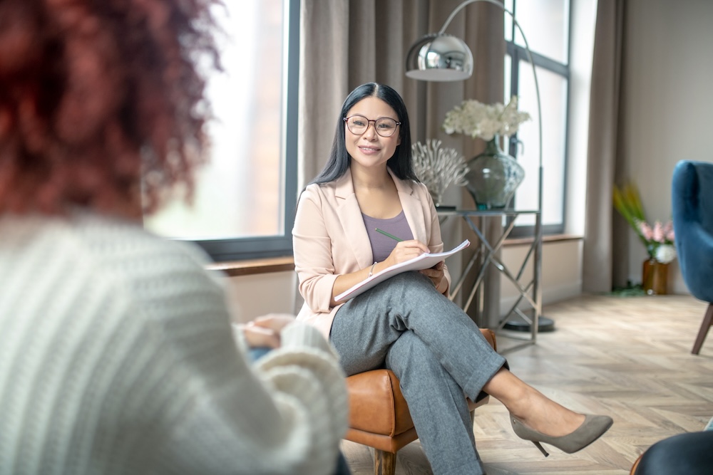 Friendly psychologist wearing grey trousers smiling to client while speaking with her