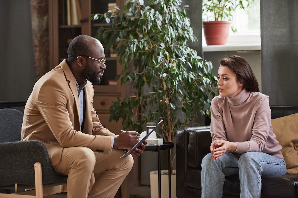 Young psychologist in beige suit making notes in clipboard while working with patient