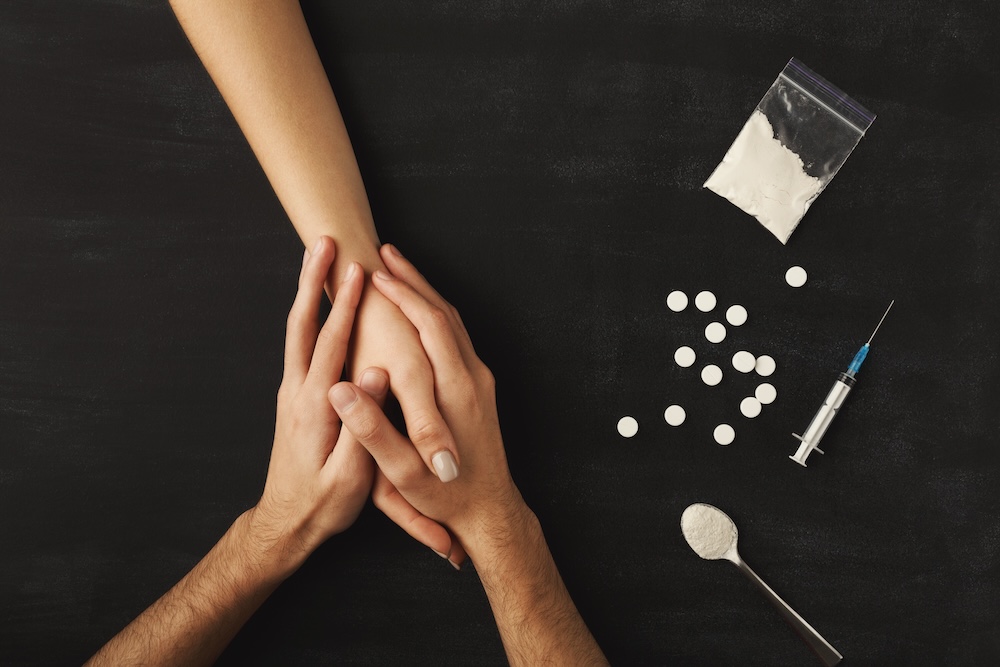 Hands of opioid addict with pills, heroin, and spoon on dark table