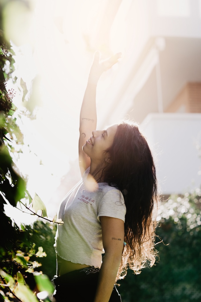 Side view of charming young female smiling and waving hand while standing near bushes on backyard