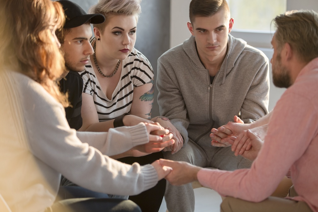 Group of young adults in alcohol and drug rehab sitting in circle, talking with psychotherapist