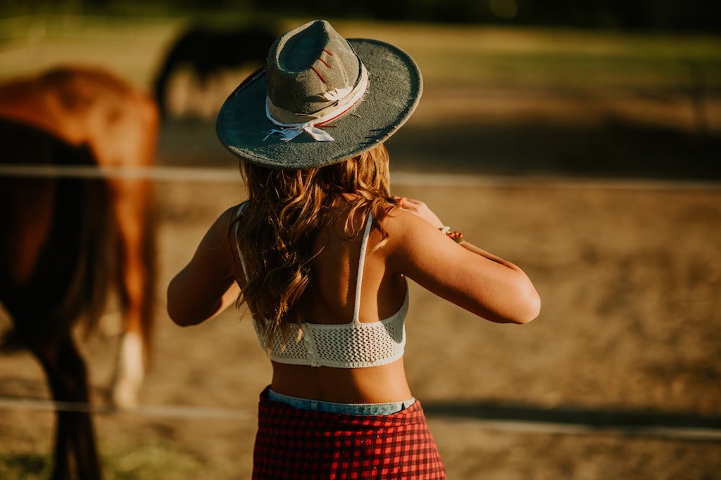 Back view of a female in cowboy hat and dressed in western styled clothes standing near fenced horse pasture with horses grazing