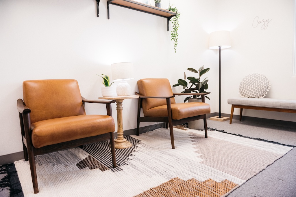 Brown chairs with lamps and house plants with decorations on a white wall in therapy office.