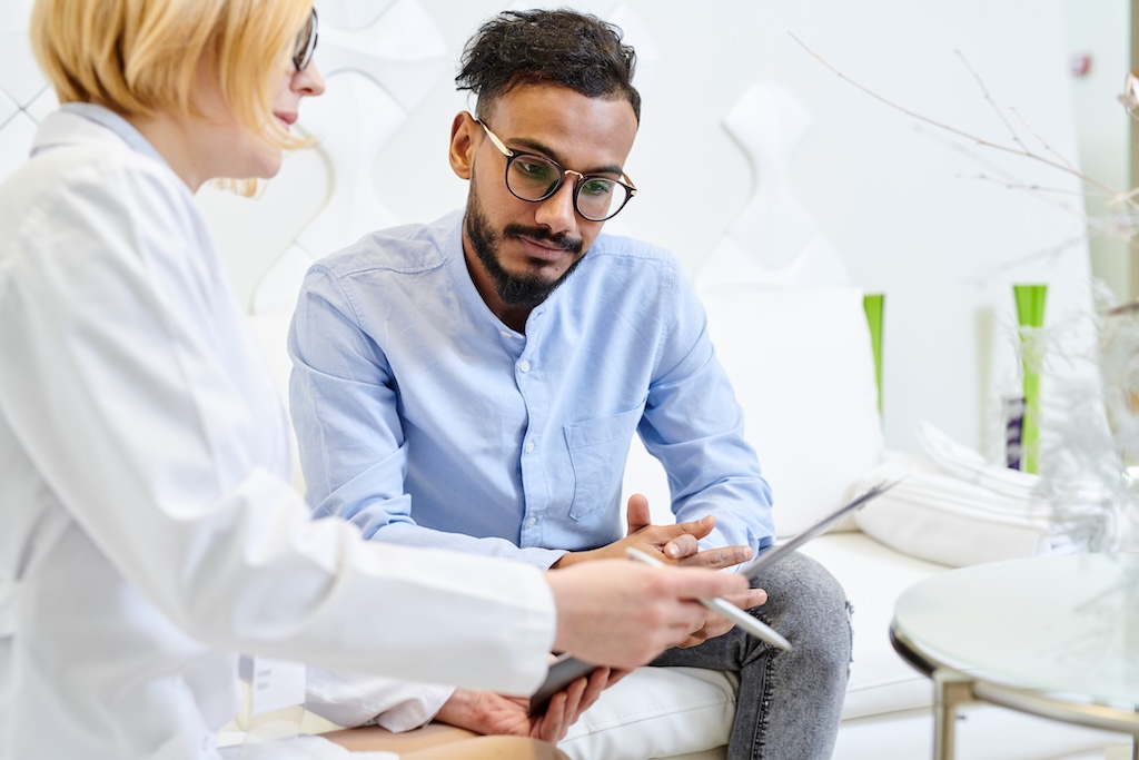 Patient answering questions of female physician while she filling in medical record, interior of modern office on background